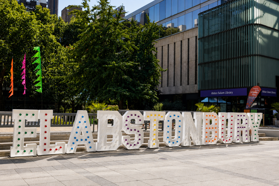 Large outdoor letter sign spelling ELABSTONBURY with multicolored dotted letters on a paved plaza, with trees, buildings, and flags in the background.