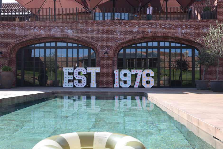 Outdoor brick building with large arched glass windows, a swimming pool in the foreground, and oversized white marquee letters spelling “EST 1976” on the pool deck.
