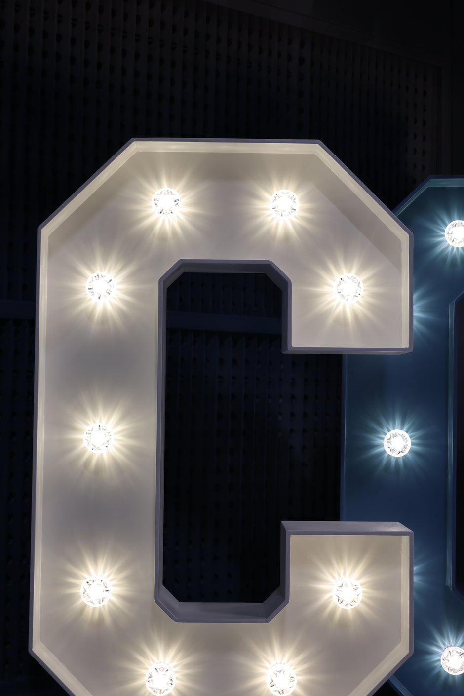 A large illuminated marquee-style letter C with warm white light bulbs, shown against a dark perforated background with part of a blue letter visible at the right edge.