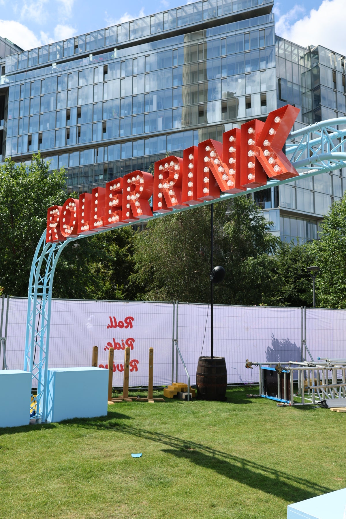A large red marquee-style sign reading "ROLLERRINK" with light bulbs is mounted on a light blue metal arch over a grassy area. A white fence, trees, and a modern glass building are visible in the background.