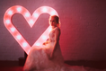 Load image into Gallery viewer, Woman in a white dress standing in front of a heart-shaped light display against a brick wall.
