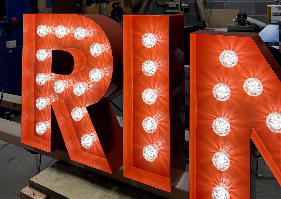 Large illuminated marquee letters spelling part of a word in bright orange-red, each filled with round light bulbs, photographed in a workshop with a gray brick wall and industrial equipment in the background.