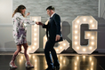 Load image into Gallery viewer, Man and woman dancing in front of large illuminated letters spelling 'JLG' in a formal setting.
