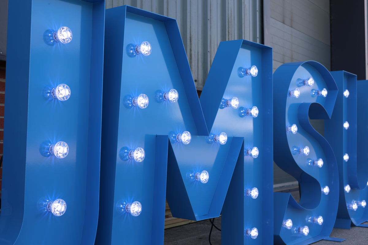 Large blue illuminated marquee letters spelling part of a word, with glowing bulb lights set into the front, photographed indoors against a gray corrugated wall.