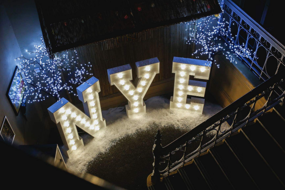 Decorative lights spelling 'NYE' on a staircase with festive lights in the background.