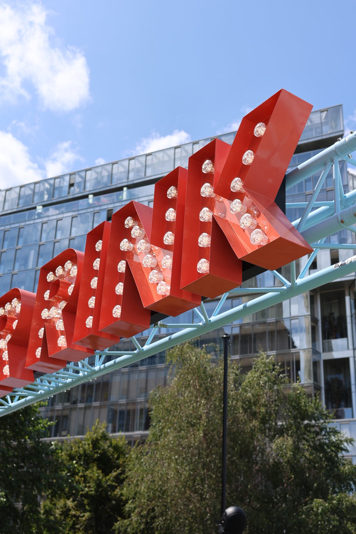 Large red illuminated sign reading "BANK" mounted on a light blue metal truss structure outdoors, with a modern glass building, blue sky, and trees in the background.