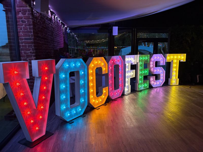 Large marquee-style letters spelling VOCOFEST arranged on a wooden floor in an indoor venue at night, each filled with multicolored LED lights.