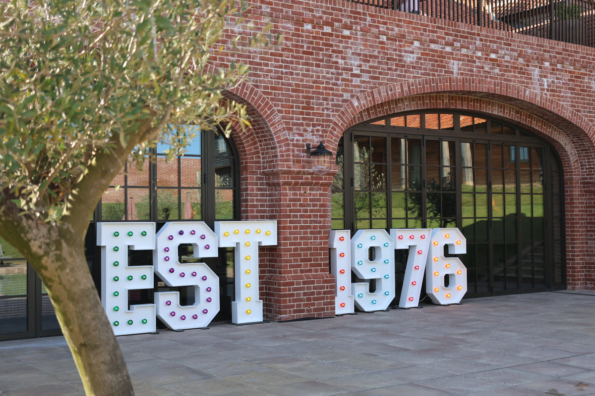 Large decorative freestanding letters spelling EST 1976 with colorful dot accents, placed outdoors in front of a red brick building with arched windows and a tree in the foreground.