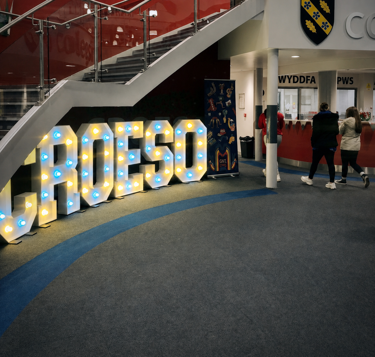 Large illuminated marquee letters spelling 'GROESO' on the floor of a modern indoor campus or hallway, with a staircase on the left and people walking in the background.