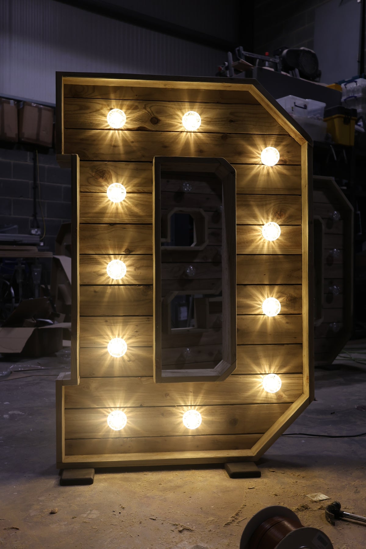 A large wooden letter 'D' marquee sign with warm glowing light bulbs, photographed in a workshop setting.